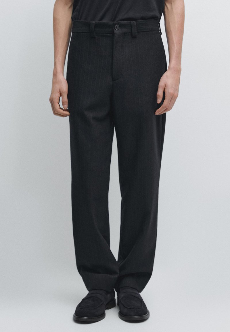 Pantalon noir à fines rayures avec une coupe structurée, un devant plat et des passants de ceinture, associé à des mocassins noirs sur un fond clair.