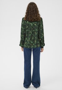 Green patterned blouse with long sleeves, cinched cuffs, and floral accents; paired with wide-leg blue jeans.
