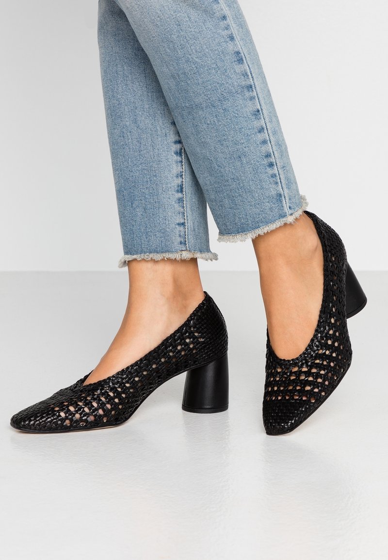 Chaussures à talons hauts noires en tressé avec un design ouvert, bout pointu et talon épais. Assorties à un jean en denim bleu effiloché coupé.