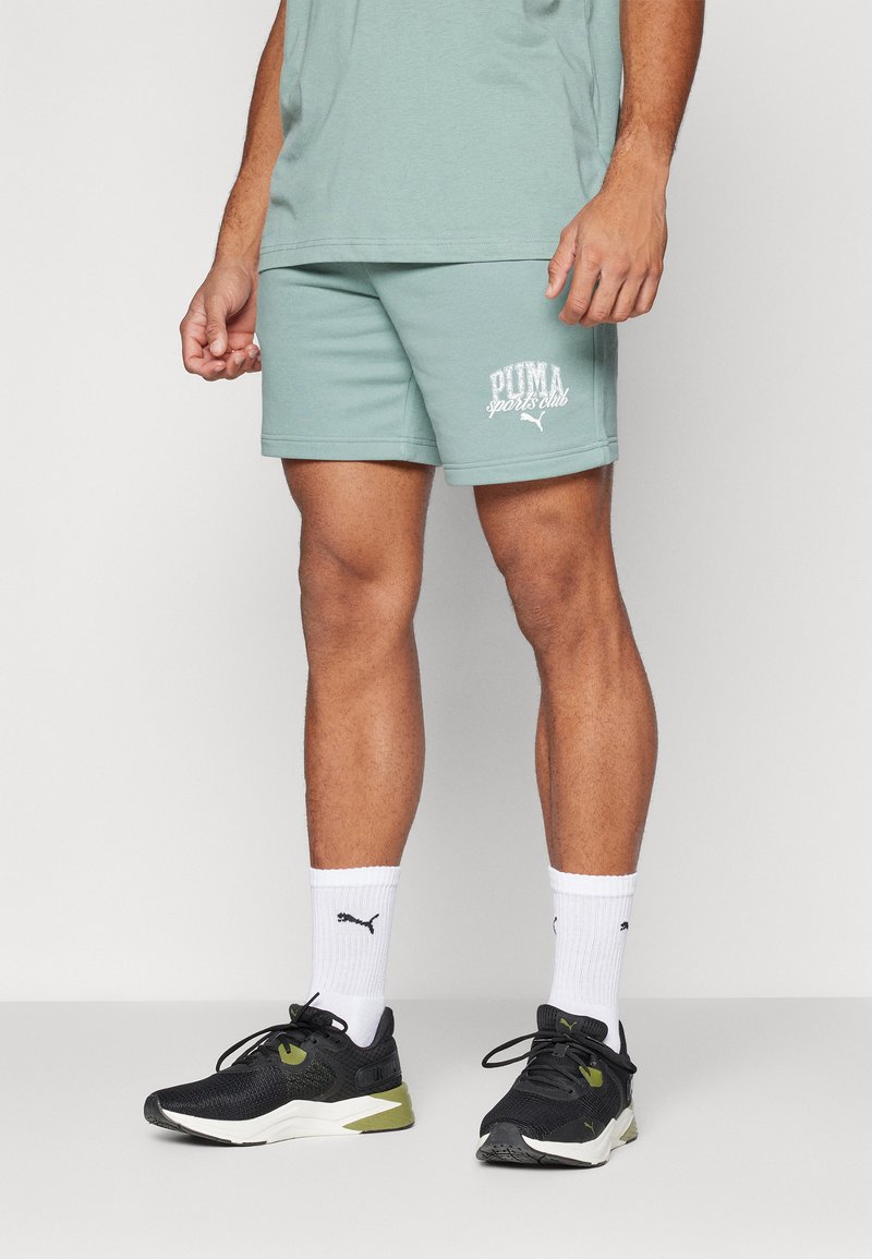 Pantalones cortos de algodón verde claro con el texto en blanco "PUMA sports club", combinados con zapatillas deportivas negras y calcetines blancos con un pequeño logo.