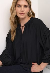 Blusa nera con design a volant e scollatura con lacci. Presenta maniche a palloncino con polsini elasticizzati e una texture morbida.