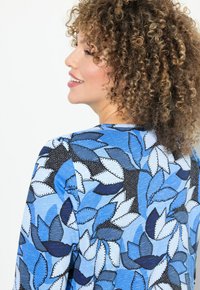 Bluse mit floralem Muster, die große Blätter in verschiedenen Blautönen zeigt, mit schwarzen Akzenten und aus einem weichen, strukturierten Stoff gefertigt.