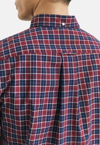 Mann trägt ein kariertes Hemd in Rot und Marineblau mit Knöpfen, der Blick ist auf den Rücken und den Kragenbereich gerichtet.