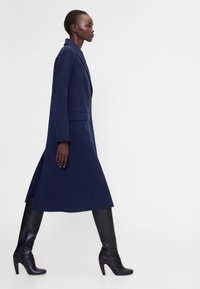 Tengerészkék dupla soros kabát hosszú, áramló sziluettel, fekete, térdig érő, magas sarkú csizmával párosítva. Éles hajtókákkal és oldalsó hasítékokkal rendelkezik.