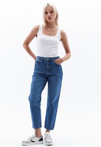 Valkohihnaiset siniset farkut, joissa on suora leikkaus, vaalea kulutus ja sivutaskut, yhdistettynä valkoiseen topiin ja mustilla yksityiskohdilla varustettuihin lenkkareihin.