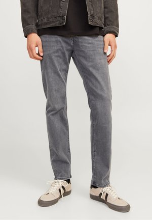 Ung voksen iført falmede grå jeans, sorte og beige sneakers og en sort denimjakke med hænder afslappet ned langs siderne.