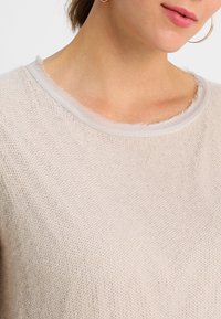 Ljus beige tröja med ett texturerat stickmönster, med en fransig halsringning och ett mjukt, fint tyg. Enkel, rundad kragsdesign.