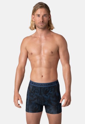 Shirtless man with medium-length blond hair stands facing forward wearing dark blue patterned boxer briefs with "MUCHACHOMALO" waistband.