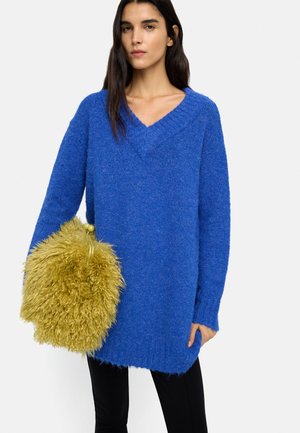 Kobieta ubrana w jasnoniebieski, za duży sweter z dekoltem w serek, trzymająca dużą, puszystą żółtą torbę na jednolitym białym tle.