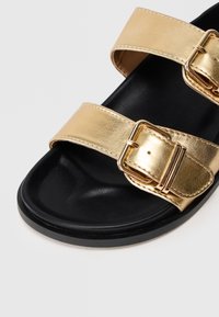 Χρυσή μεταλλική σαγιονάρα με δύο φαρδιές, ρυθμιζόμενες λουρίδες που διαθέτουν κούμπωμα. Μαύρη σόλα με απαλό, υφασμένο πέλμα.