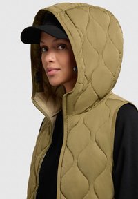 Gilet trapuntato verde oliva con cappuccio oversize, chiusura a zip e sottile motivo a onde; indossato sopra una maglietta nera, completato da un cappellino nero.
