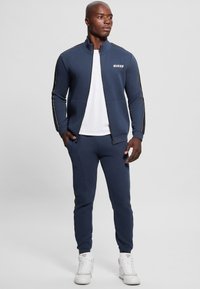 Completo sportivo blu navy composto da una giacca con zip e logo, strisce laterali e pantaloni abbinati. Indossato con una maglietta bianca e sneakers bianche.