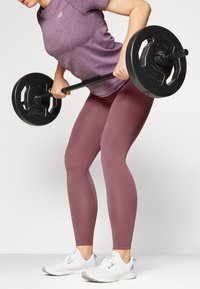 Un haut de sport violet, des leggings maroon ajustés et des chaussures de sport blanches. La personne soulève une barre noire avec des poids ronds.