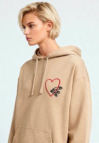 Sweatshirt beige avec un grand motif de cœur rouge et des épingles de sécurité noires brodées sur la poitrine. Il comprend des cordons de serrage et une poche frontale.