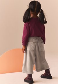 Fată îmbrăcată cu un tricou lung de culoare vinom și pantaloni gri deschis cu dungi și buzunare. Poartă bocanci negri cu talpă groasă și are două codițe.
