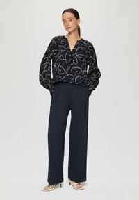 Blouse noire avec un motif de cœurs blancs, décolleté en V et manches bouffantes, associée à un pantalon large bleu marine et des talons blancs à lanières.