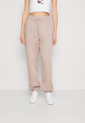 Persona que lleva pantalones de chándal beige de talle alto con cordón y zapatillas blancas, de pie contra un fondo liso y claro.