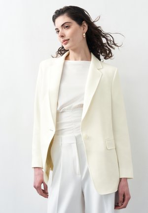 Kvinde med bølget hår iført en cremefarvet blazer, hvid top og hvide bukser med høj talje foran en ensartet lys baggrund.