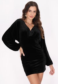 Rochie mini din catifea neagră cu decolteu adânc în V, mâneci lungi și voluminoase și o siluetă ajustată. Textură netedă cu un luciu subtil. Colier din lanț auriu.