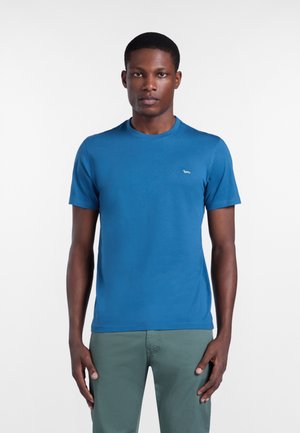 Muž oblečený v jednofarebnom modrom tričku s krátkym rukávom a zelených nohaviciach stojaci na jednoduchom bielom pozadí.