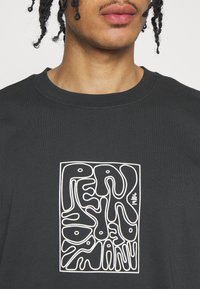 Jeune personne aux cheveux tressés portant un T-shirt noir avec un texte abstrait ondulé blanc et l’année « 1986 » au centre de la poitrine.