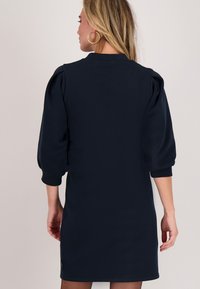 Marineblauwe jurk met pofmouwen, ronde hals en een aansluitend ontwerp. Soepele stof met een subtiele textuur; heeft een zichtbaar zijlabel.