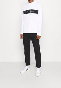 Witte hoodie met een marineblauwe middenstrook, voorzien van tekst en logo; gecombineerd met zwarte slim-fit broek en witte sneakers met zwarte accenten.
