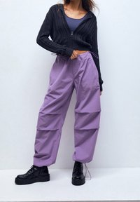 Hoodie noir en tricot court avec motif en torsades, associé à un pantalon ample lavande avec poches et cordon de serrage ajustable, porté avec des bottes noires.