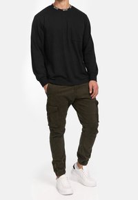 Camicia nera a maniche lunghe con taschino sul petto e collo con logo, abbinata a pantaloni cargo verde oliva con molte tasche e polsini elasticizzati.