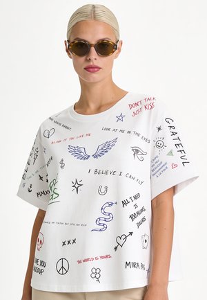 Blonde vrouw met ronde zonnebril en wit T-shirt met kleurrijke handgeschreven zinnen en kleine tekeningen, staand tegen een witte achtergrond.