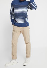 Blå stickad tröja med vita horisontella ränder, beige byxor och vita sneakers. Tröjan har rund hals och ribbade ärmslut.