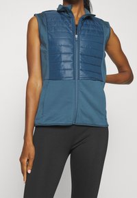 Ärmlös blå väst med en vadderad framsida i quiltat tyg och släta sidor i tyg. Har dragkedjestängning och uppstående krage, utformad för aktiv användning.