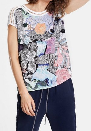 Mujer que lleva una camiseta de manga corta con ribete de crochet y un estampado colorido de peces, flores y formas abstractas, combinada con pantalones de color azul marino con cordón.