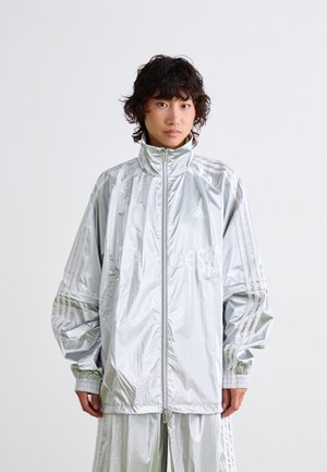 Personne avec des cheveux courts et bouclés portant une veste zippée Adidas réfléchissante en argent et un pantalon assorti sur fond blanc uni.