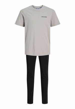 Blond boy standing barefoot wearing a light grey short-sleeve shirt and black pants against a plain white background.