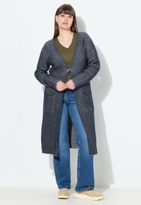 Pitkä, tummansininen kardiigan, jossa on ribbitekstuuri, avoin etuosa, kaksi etutaskua ja hieman ylisuurennettu malli, päällä vihreä paita ja farkut.