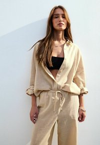 Beige lättviktslinne skjorta, knäppt uppe, ihop med matchande löst sittande byxor som har resårmidja och dragsko.