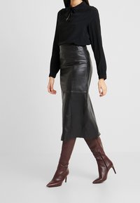 Zwarte blouse met lange mouwen en strikdetail, gecombineerd met een zwart leren midi-rok met hoge taille en puntige bordeauxrode kniehoge laarzen.