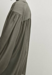 Long-sleeve shirt in olive green fabric, featuring a gathered back design and relaxed fit. Soft texture with a subtle sheen.