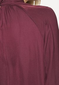 Blusa borgogna con collo alto, texture morbida e dettagli arricciati sul retro, che mette in risalto una silhouette fluida.