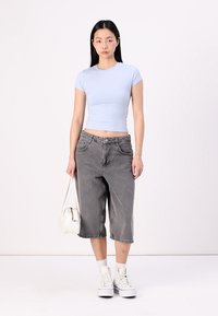 T-shirt ajustada de cor azul clara, calças de ganga largas e cortadas cinzas, sapatilhas brancas de cano alto e uma pequena mala branca, apresentadas contra um fundo simples.