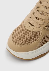 Μπεζ αθλητικά παπούτσια με επάνω μέρος από δίχτυ, υφή στην επιφάνεια, στρογγυλή μύτη και λευκή σόλα μέσης με εξωτερική σόλα από καουτσούκ γκουμ. Ορατές λεπτομέρειες με ραφή.