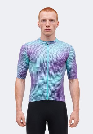 Homme aux cheveux roux courts portant un maillot de cyclisme moulant bleu et violet dégradé avec une fermeture éclair sur le devant et un cuissard noir.