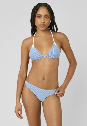 Femme aux cheveux tressés portant un bikini rayé bleu et blanc avec des bretelles en corde nouées autour du cou, debout devant un fond gris uni.