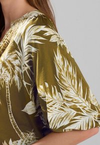 Olive green blouse featuring white tropical leaf print, short sleeves, and a relaxed fit. Soft fabric with subtle sheen and gathered neckline.