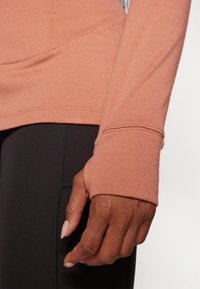 Camisola de manga comprida em tecido rosa suave e empoeirado, com buracos para polegares e uma bainha arredondada, combinada com leggings de compressão pretas.