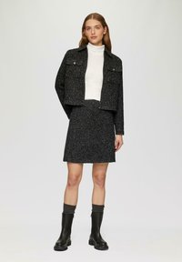 Schwarze strukturierte Jacke mit cropped Schnitt, kombiniert mit einem weißen Rollkragenpullover und einem passenden schwarzen Rock. Getragen mit kniehohen schwarzen Stiefeln und grauen Socken.