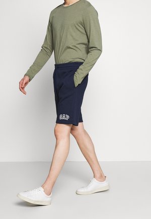 Pantaloni corti blu navy con logo "GAP" in bianco, realizzati in tessuto morbido. Abbinati a una camicia a maniche lunghe verde oliva e scarpe da ginnastica bianche.