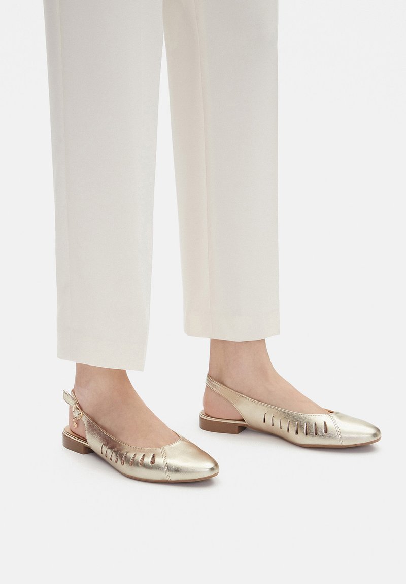 Zapatos planos slingback metálicos dorados con recortes abiertos. Punta redondeada, tacón bajo de bloque y correa slingback con detalle de hebilla ajustable.