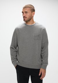 Grå sweatshirt i texturerat tyg med rund halsringning, långa ärmar och en subtil bröstlogotyp. Materialet ser mjukt och avslappnat ut.